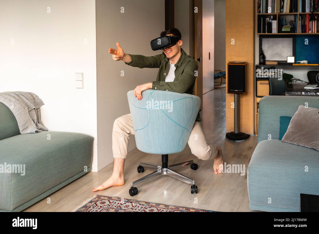 Smiling young man wearing virtual reality simulator gesturing on chair at home Stock Photo