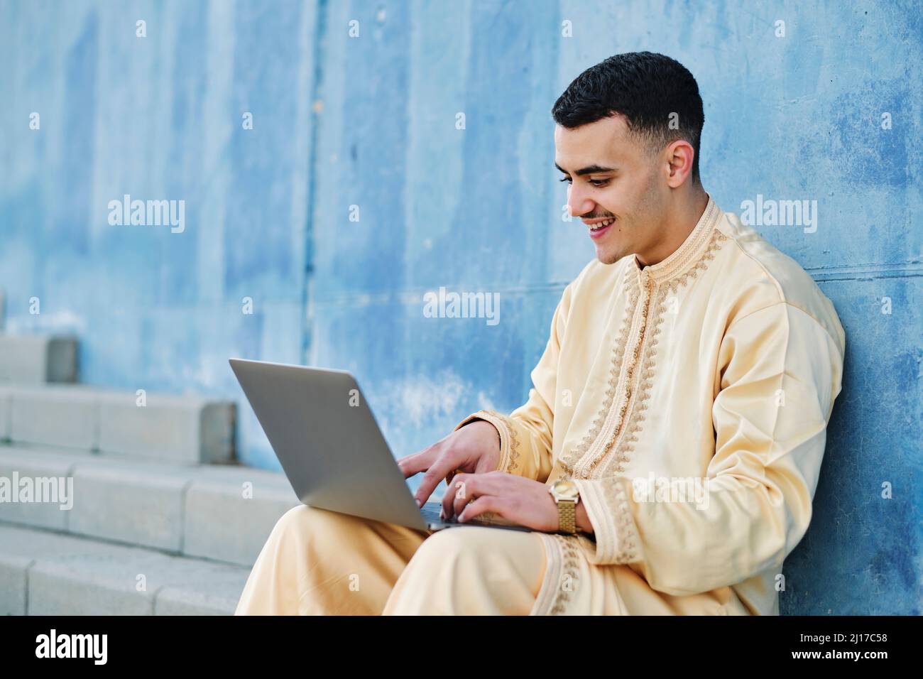 Happy young man using laptop sitting on steps by blue wall Stock Photo