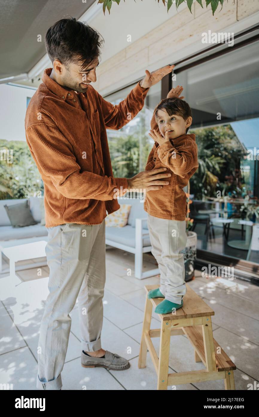 Father measuring height of son standing on stool in patio Stock Photo