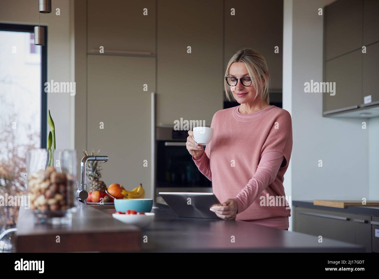Blond woman holding coffee cup using tablet PC in kitchen Stock Photo