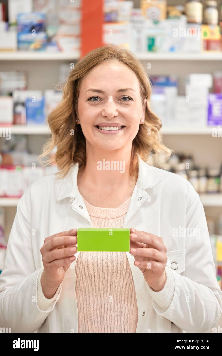 Happy female pharmacist holding green box of medicine at pharmacy store Stock Photo