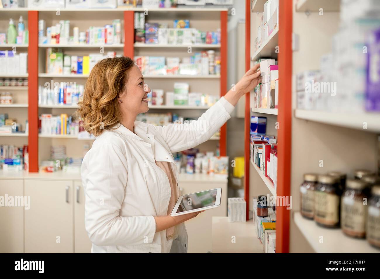 Smiling pharmacist holding tablet PC stacking medicine in shelf at pharmacy store Stock Photo