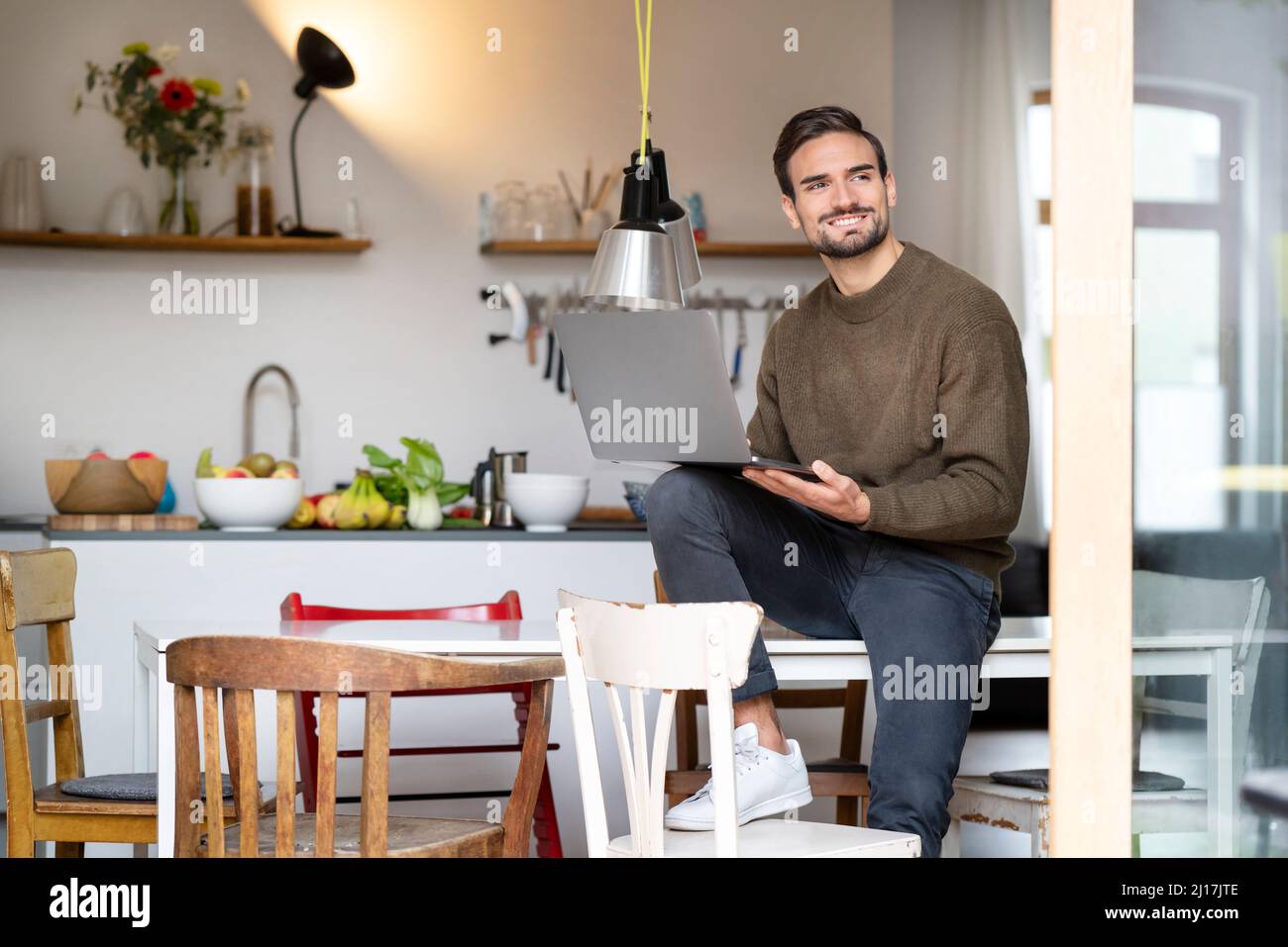 Smiling freelancer with laptop sitting on table in kitchen at home ...
