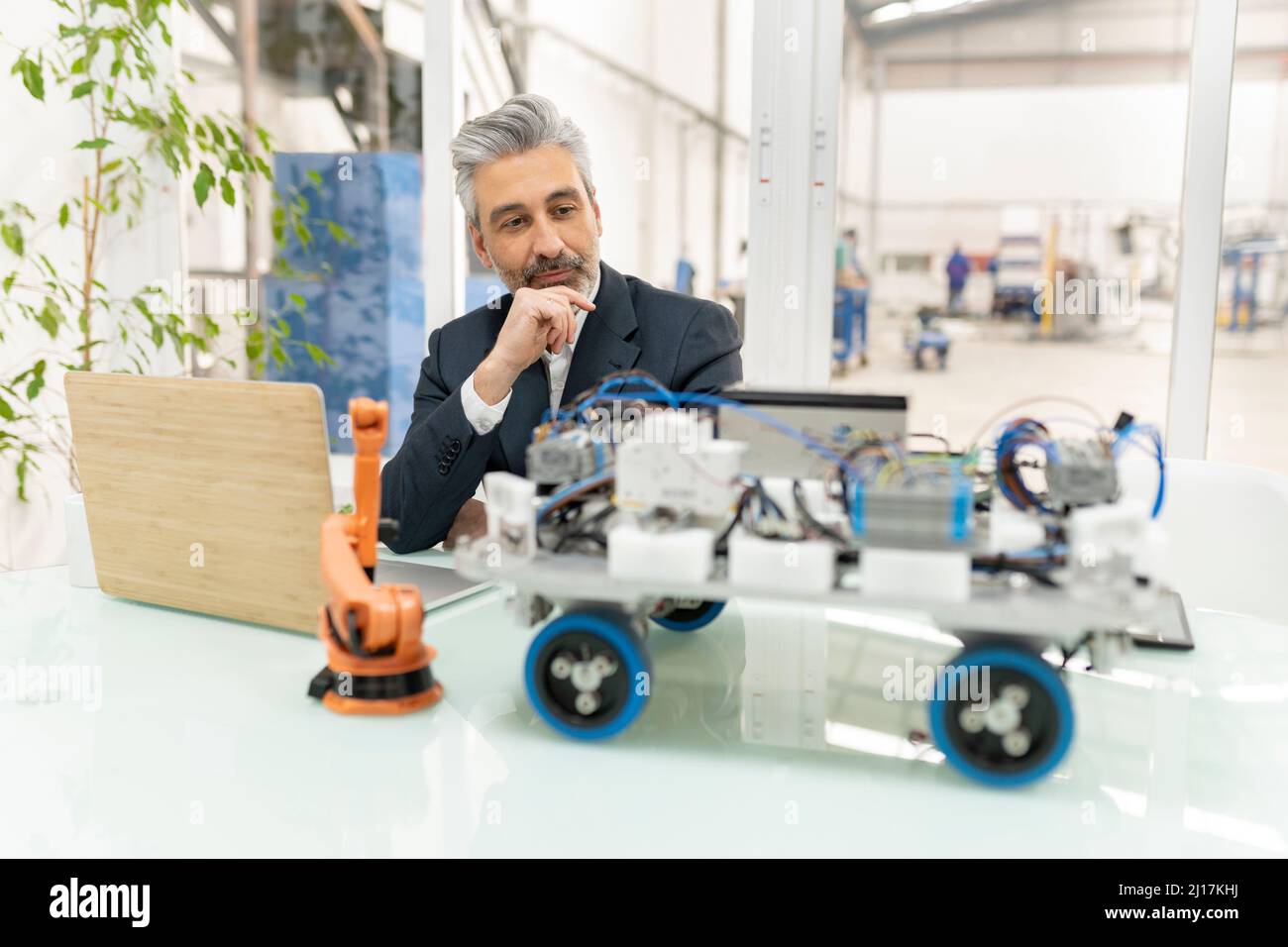 Businessman sitting with hand on chin looking at model of robotic vehicle in factory Stock Photo