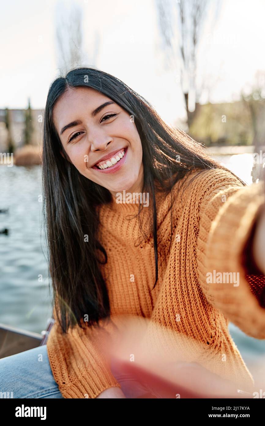 Photo of carefree long haired young latin woman takes selfie portrait on mobile phone, wears yellow colorful jumper, uses technology during leisure ti Stock Photo