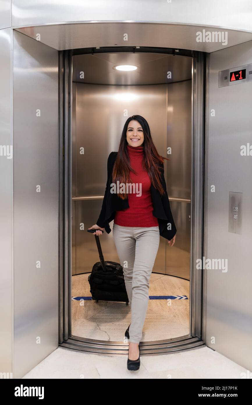 Happy working woman with wheeled luggage moving out from elevator Stock Photo