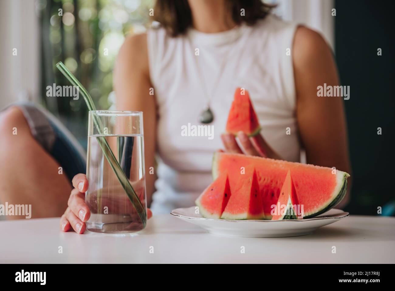 Woman holding drinking glass by watermelon slice in plate on table at home Stock Photo