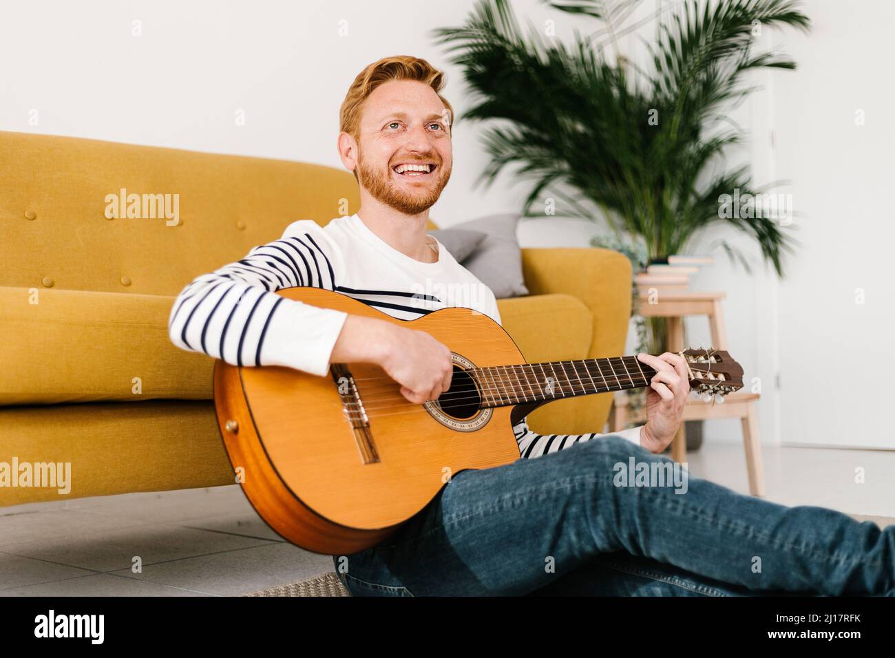 Cheerful man with guitar sitting in front of sofa at home Stock Photo
