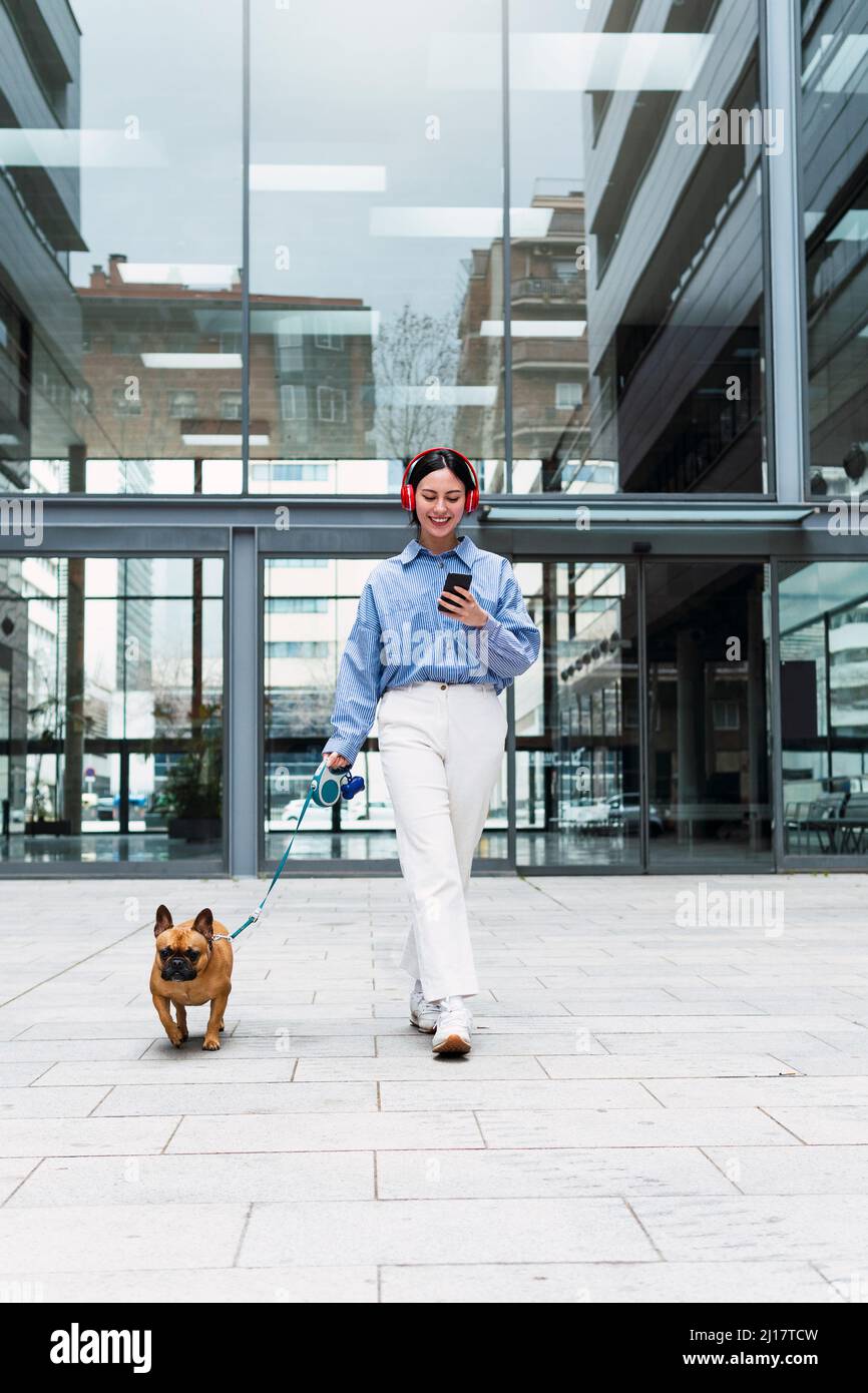 Happy woman listening music through wireless headphones walking with dog in front of building Stock Photo