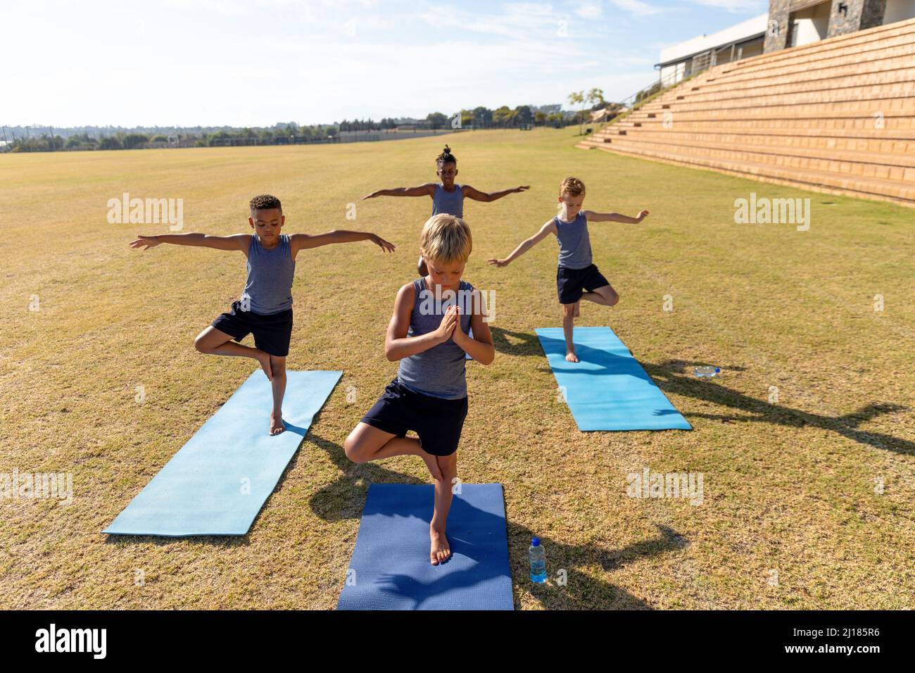 Multiracial elementary schoolboys with arms outstretched and hand clasped standing on one leg on mat Stock Photo