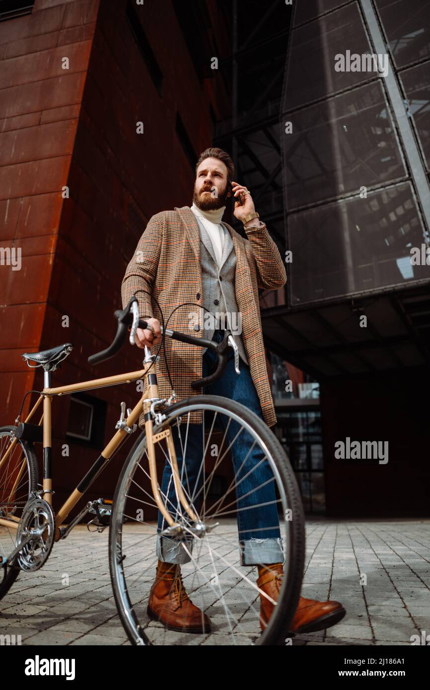 Young hipster businessman walking with his bike and talking on phone. Stylish manager in business area. Outside Stock Photo