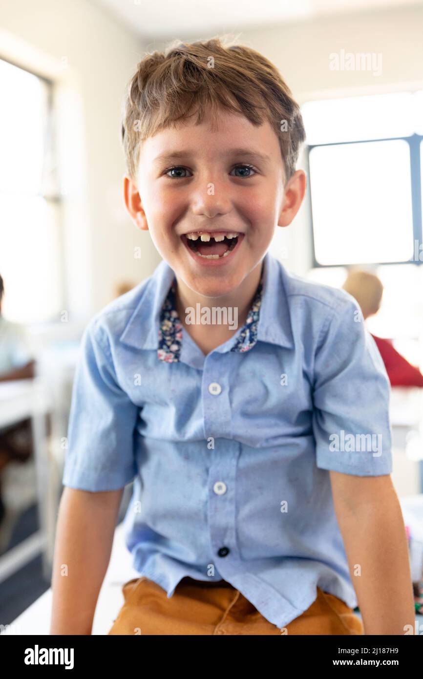 Portrait of cheerful cute caucasian elementary schoolboy sitting on desk in classroom Stock Photo