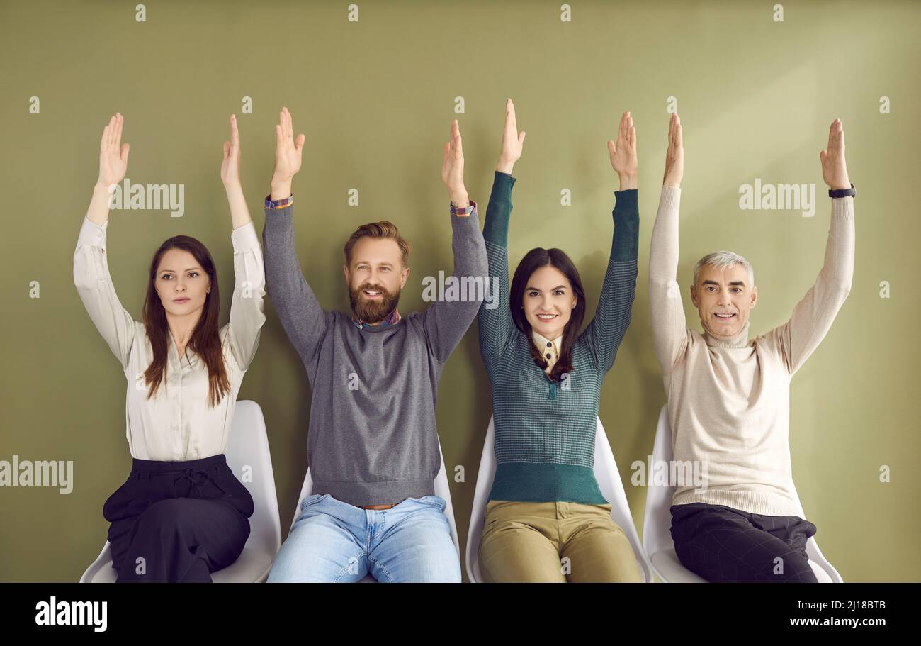 Portrait of people sitting in row on chairs and voting unanimously, raising two hands each. Stock Photo