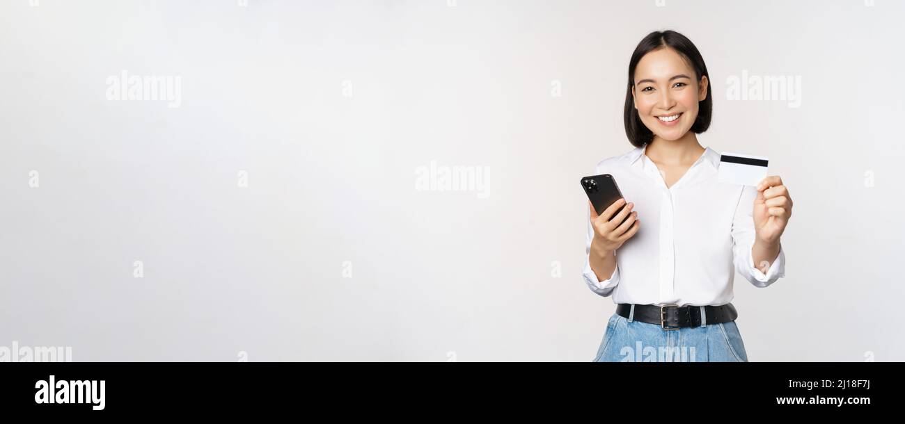 Online shopping concept. Smiling modern asian girl shows her credit card, holds mobile phone