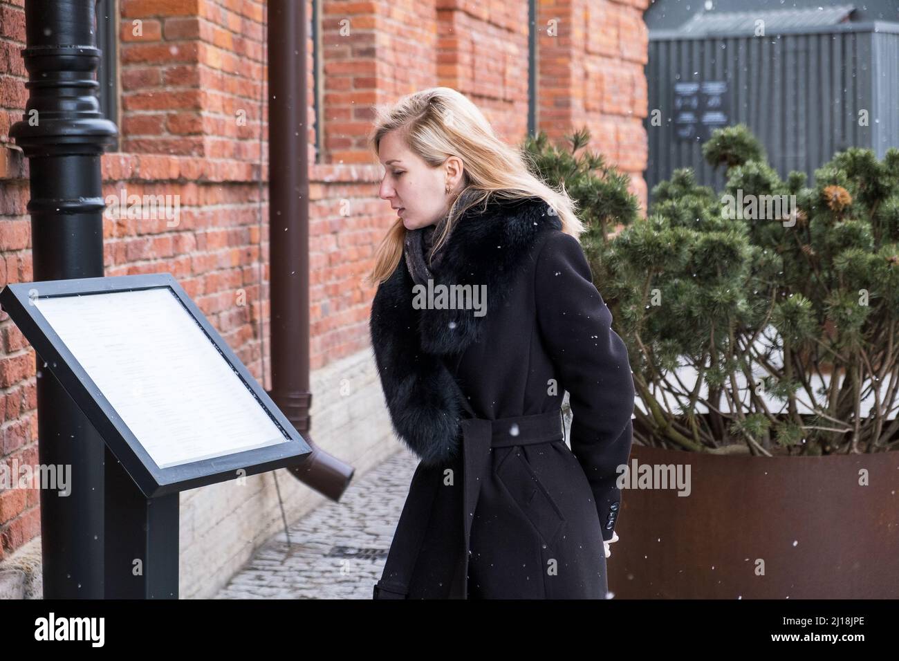 Young blonde woman in black winter coat with fur collar reads menu Stock Photo