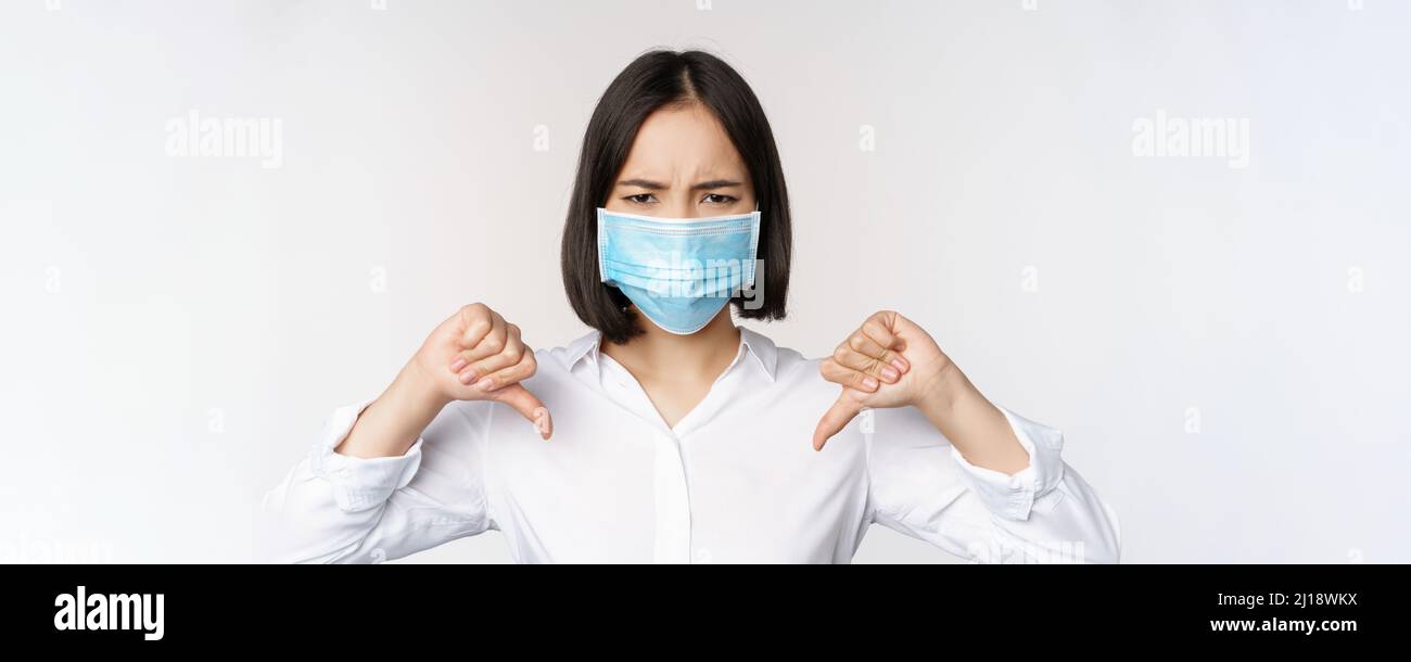 Portrait of asian woman in medical face mask showing thumbs down with ...