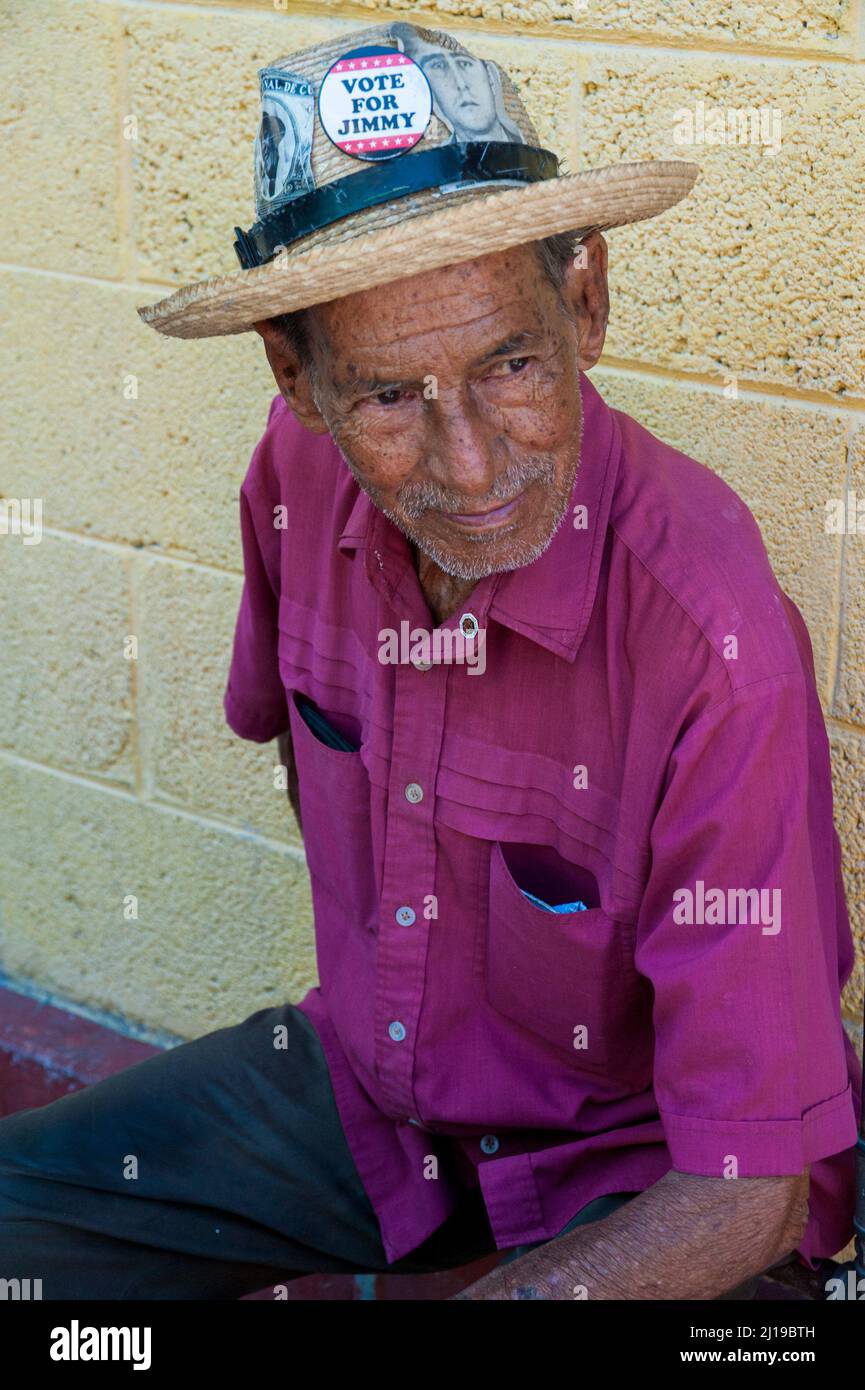 Smiling man looks around the street in Havana, Cuba while wearing a fedora hat with a vintage image of Fidel Castro and nameof President Jimmy Carter. Stock Photo