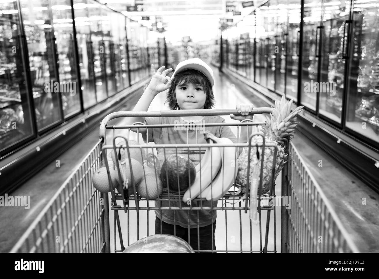 Sale, consumerism and people concept - happy little boy with food in shopping cart at grocery store. Little boy in the supermarket. Stock Photo