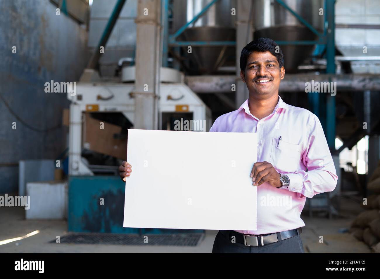smiling supervisor holding empty sign board while standing in front of machinery at factory - concept of skilled labour, job advertisement or Stock Photo