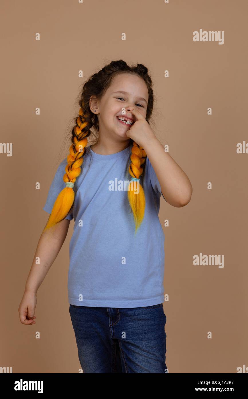 Caucasian naughty small girl smiling shining picking nose with finger looking at camera having kanekalon braids on beige background wearing blue t Stock Photo