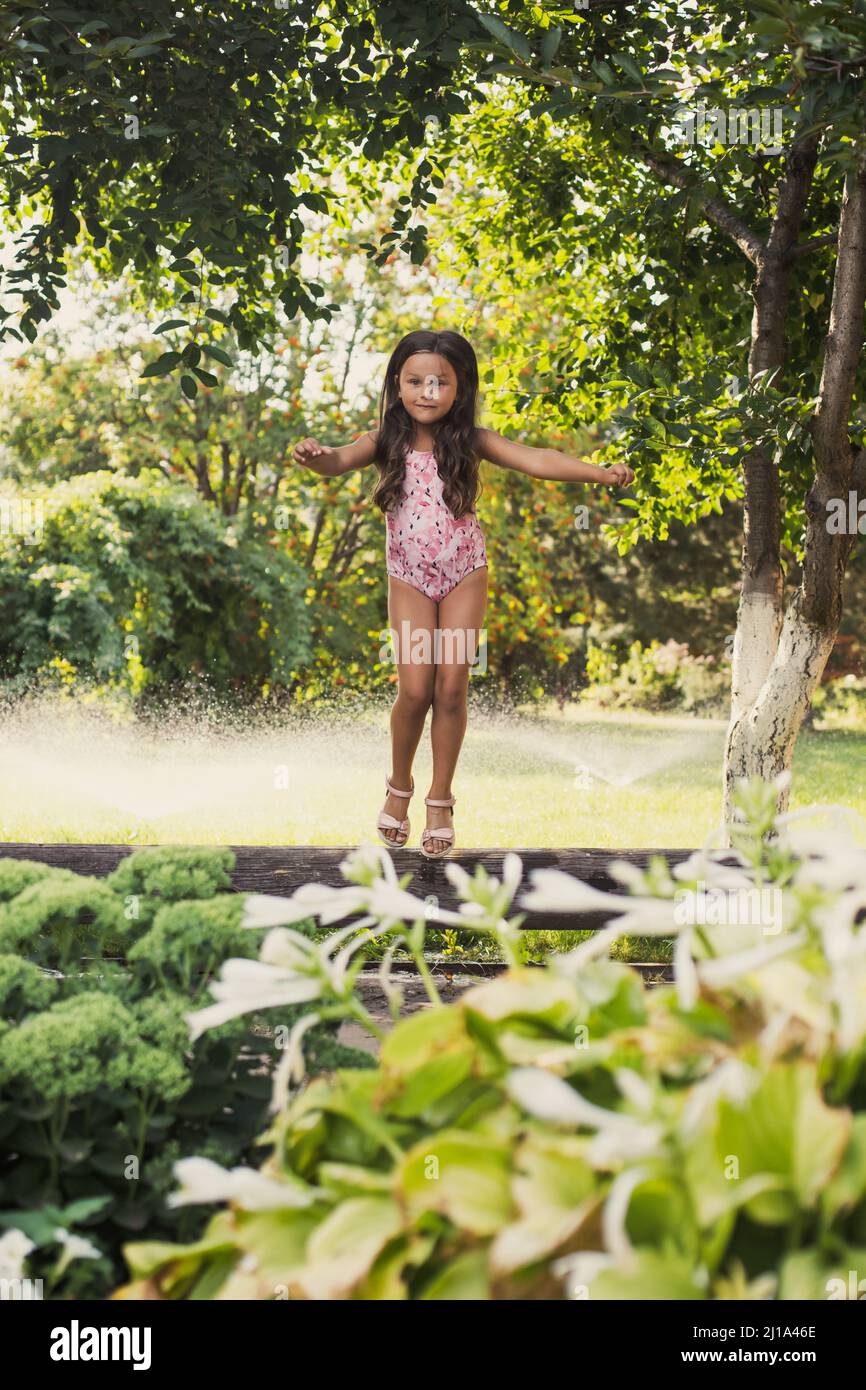 Happy young Caucasian girl balancing on log looking at camera surprisingly in backyard with green trees and water sprinklers in background in daytime Stock Photo