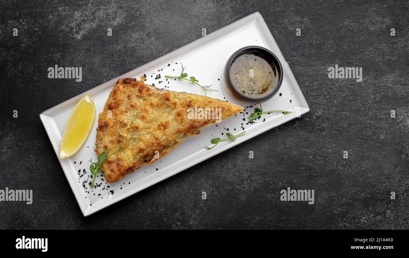 A piece of fried flounder fish with lemon on a plate, on a dark