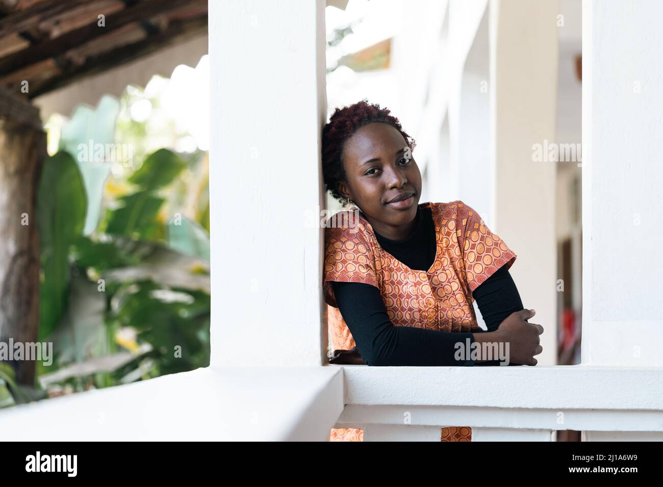 Gorgeous young African girl leaning comfortably on the balustrade of the veranda at her parent's home, smiling confidently at the camera; concept of f Stock Photo