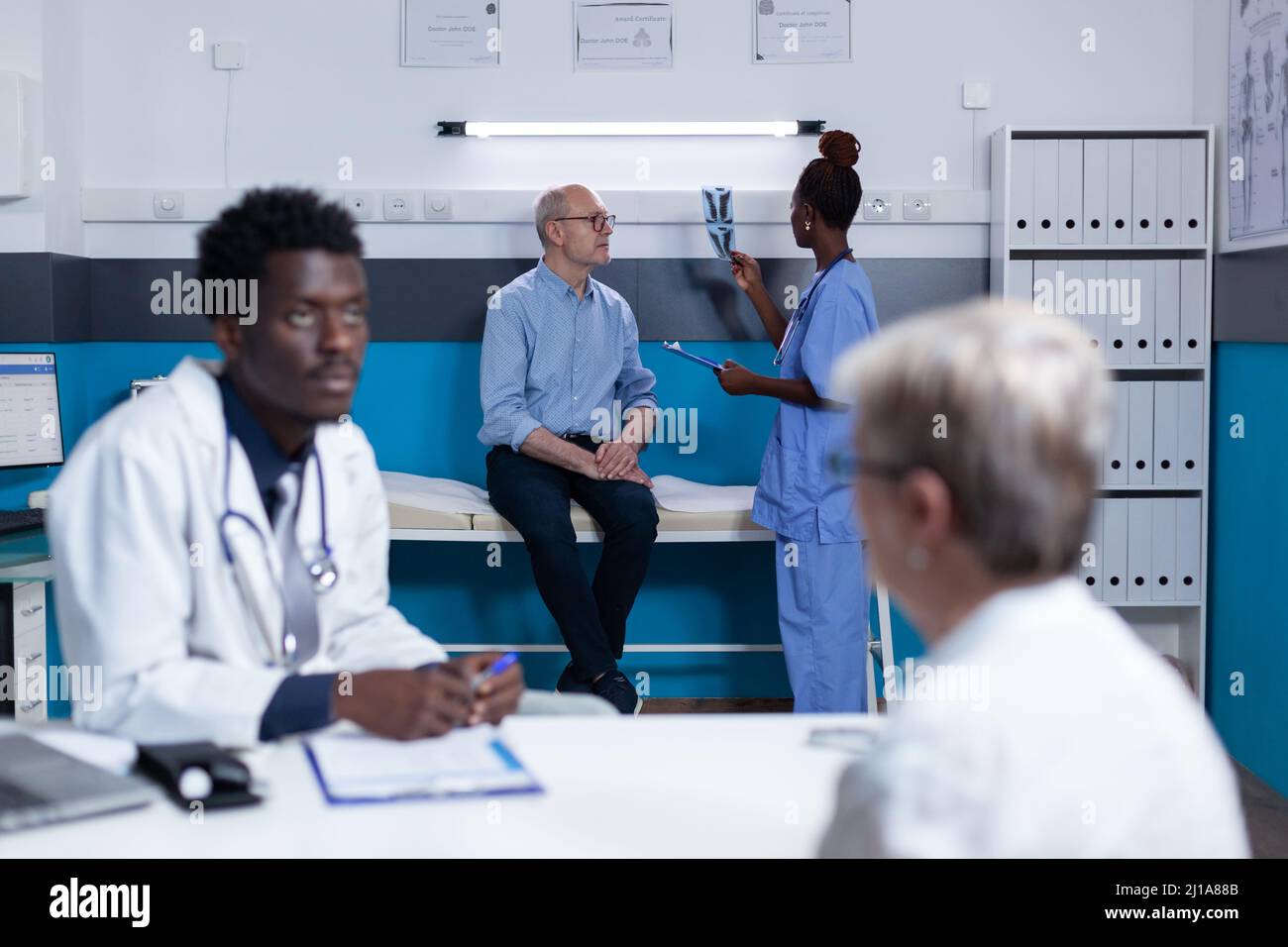 Clinic nurse verifying radiology scan results while senior patient is sitting on hospital bed. Hospital staff analyzing x-ray scan image of retired man accusing acute back pain. Stock Photo