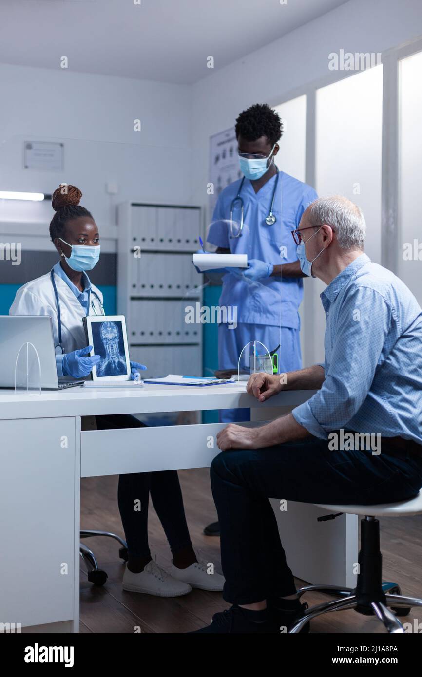 Radiology specialist holding x-ray scan image and explaining treatment schedule to retired man while medical nurse taking notes on clipboard. People wearing virus protection facemask in hospital workspace Stock Photo
