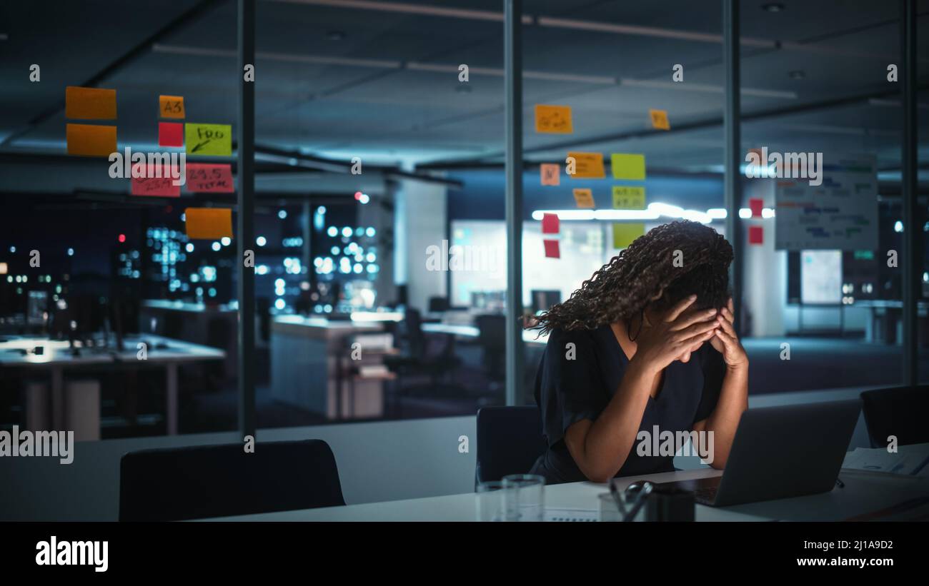 Overworked African American Businesswoman Working on Laptop Computer in Big City Office Late in the Evening. Tired Stressed Female Entrepreneur trying Stock Photo