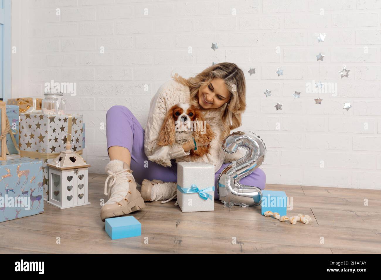 Happy well-groomed woman hug dog spaniel tight, sitting on floor near gift boxes and silver number two made of balloon. Stock Photo