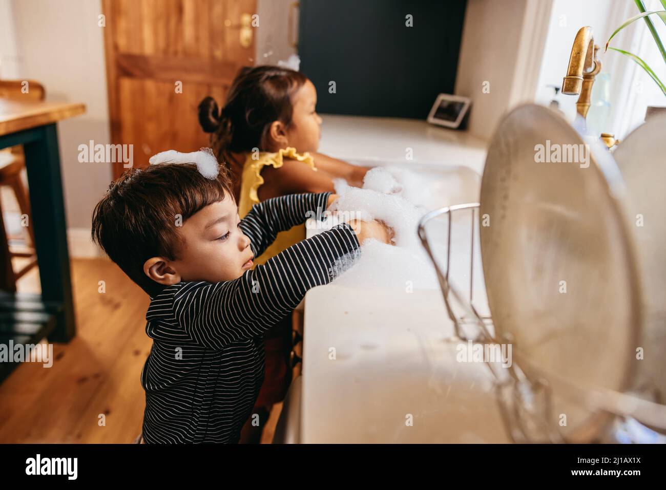 Two little toddlers playing with white foam at home. Two adorable young siblings sticking their hands into a sink full of bubbles at home. Stock Photo