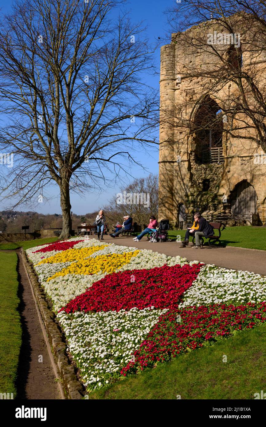 People sitting relaxing enjoying sun (bright border flowers, ancient tower keep ruins, blue sky) - Knaresborough Castle, North Yorkshire, England, UK. Stock Photo