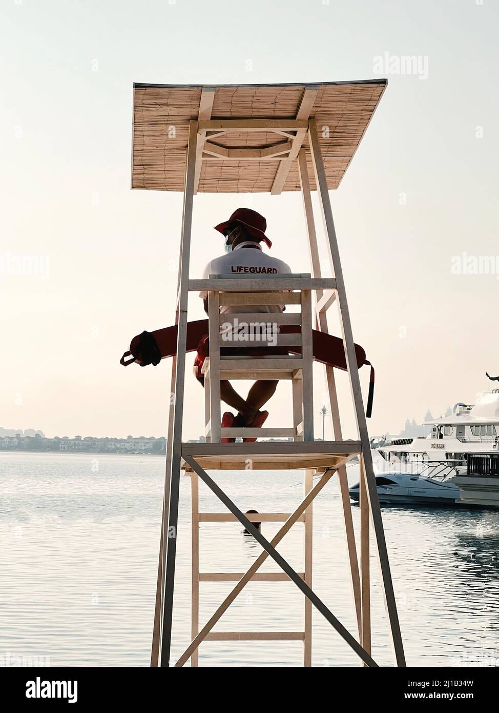 Lifeguard is sitting on wooden chair on tower watching the sea in Dubai. Boat is moored, the sea is calm. Nobody is in water Stock Photo