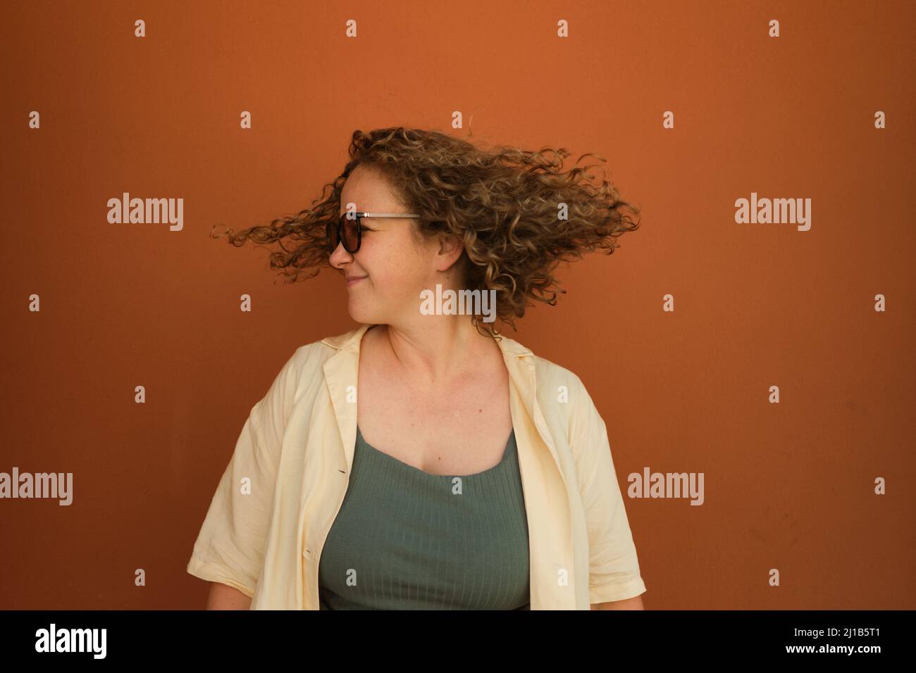Happy caucasian white girl on orange background shakes her head. Curly hair flies to sides. Hair care. Happyness. Copy space. Stock Photo