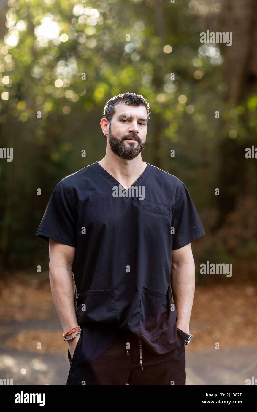 A doctor with dark hair and a beard in black scrubs standing outside in