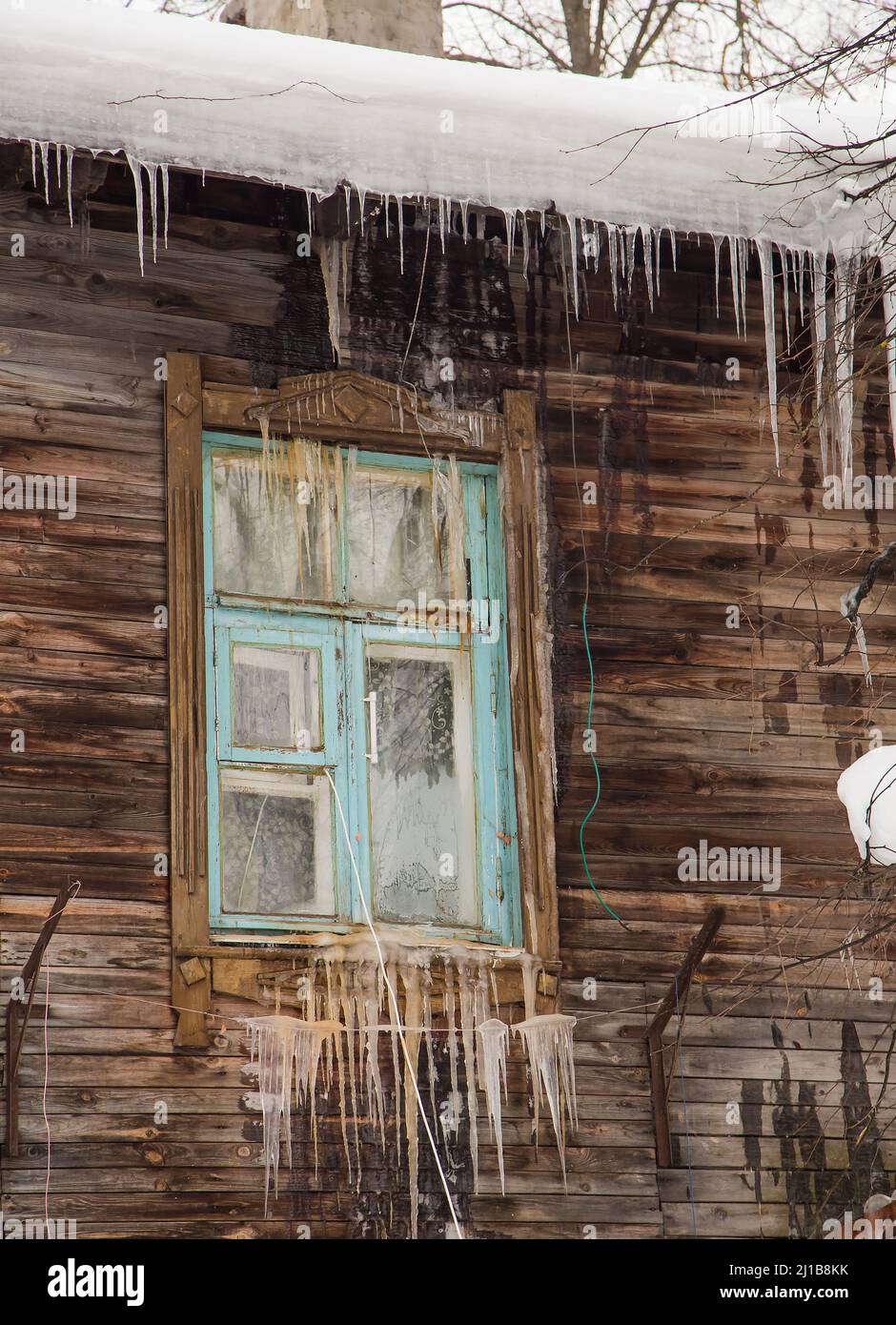 Glass transparent icicles hang on the edge of the roof and the window. Against the background of the wooden wall of the old house. Large cascades, even beautiful rows. Cloudy winter day, soft light. Stock Photo