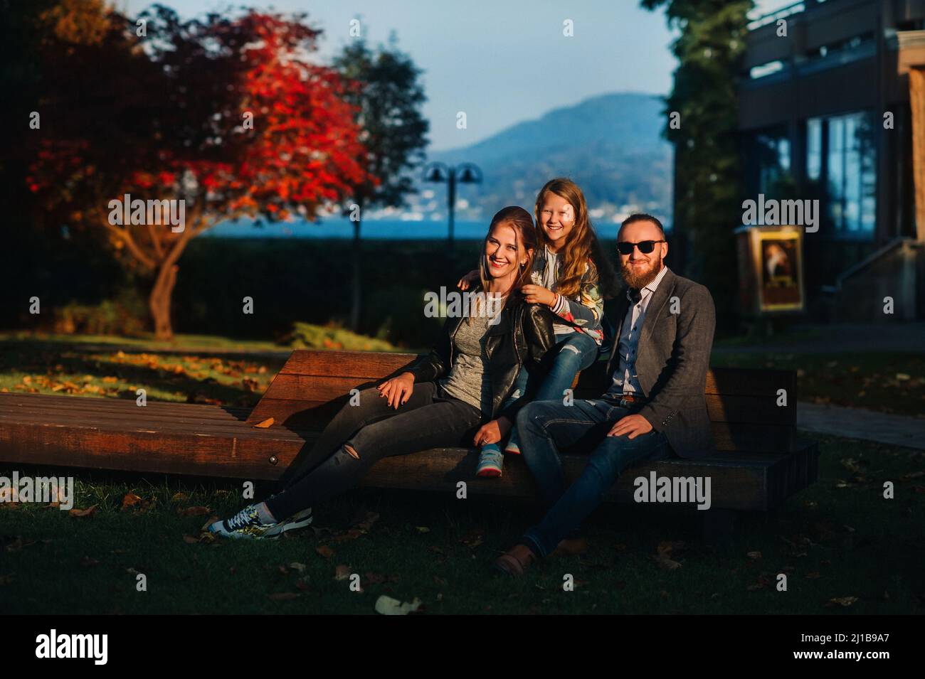 A happy family of three sits on a bench at a Sunny autumn sunset in Austria's Old town.A family poses in a small Austrian town against the backdrop of Stock Photo