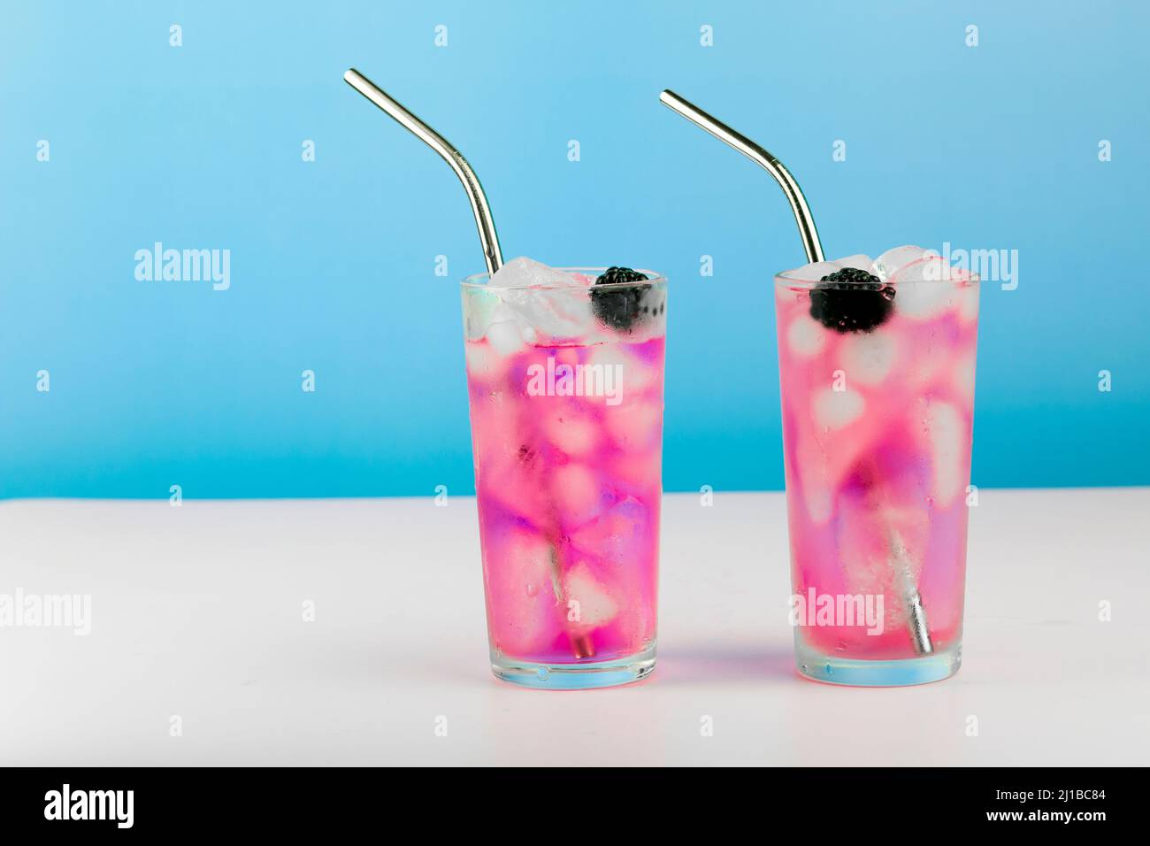 Summer refreshing pink cocktail with ice cube, metal straw and blackberry on blue background. Stock Photo