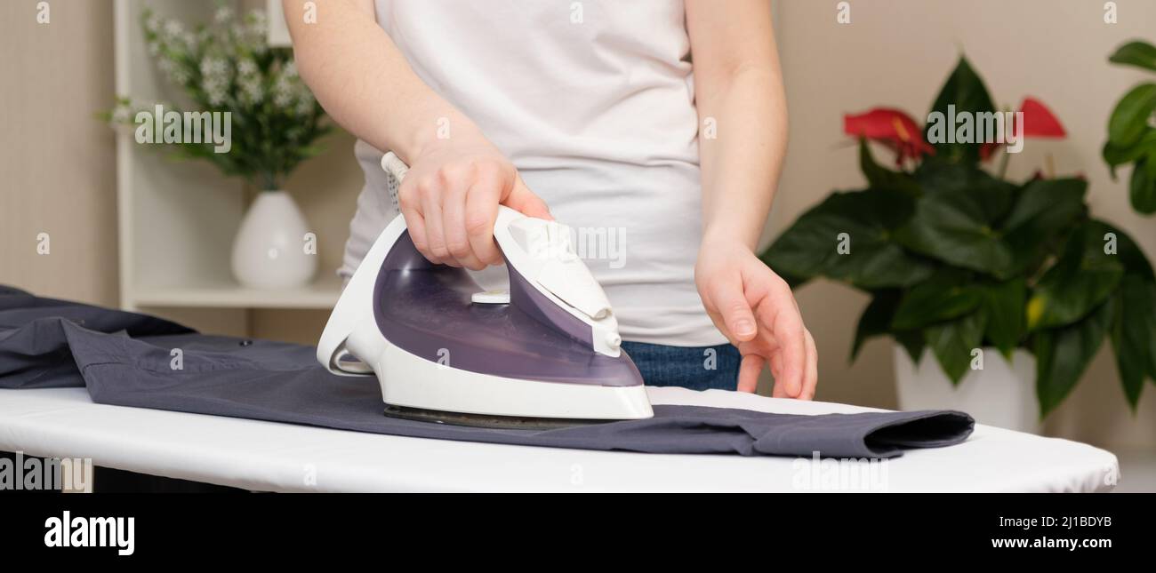 A woman irons a gray men's shirt with an iron on an ironing board at home, close-up hands. Clothing care, long banner. Stock Photo