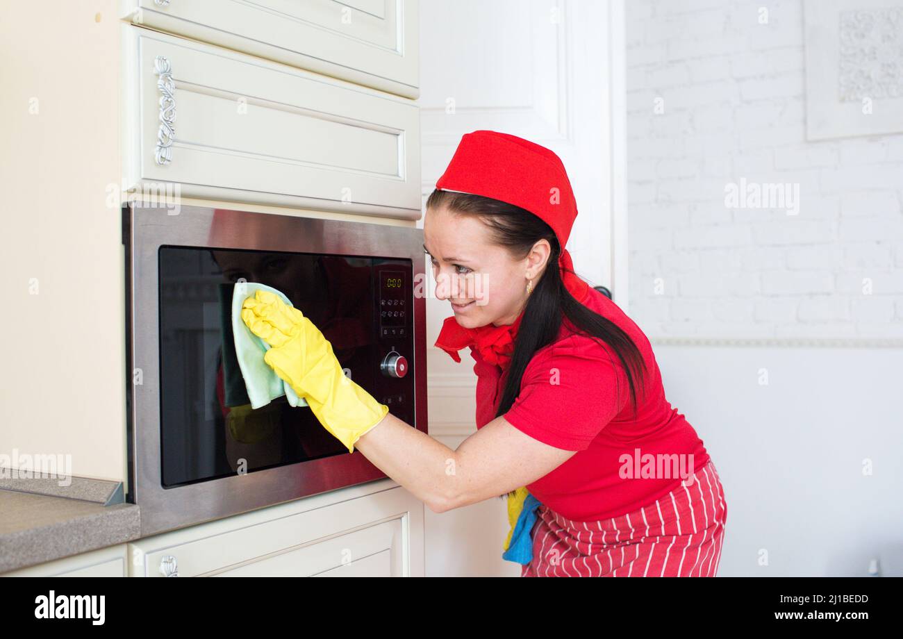 A young woman in a red uniform cleans the microwave oven with a rag. Cleaning company Stock Photo