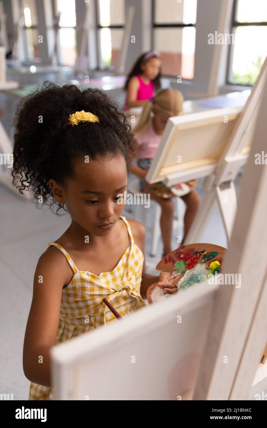 Biracial elementary schoolgirl holding paint palette while painting on easel during drawing class Stock Photo