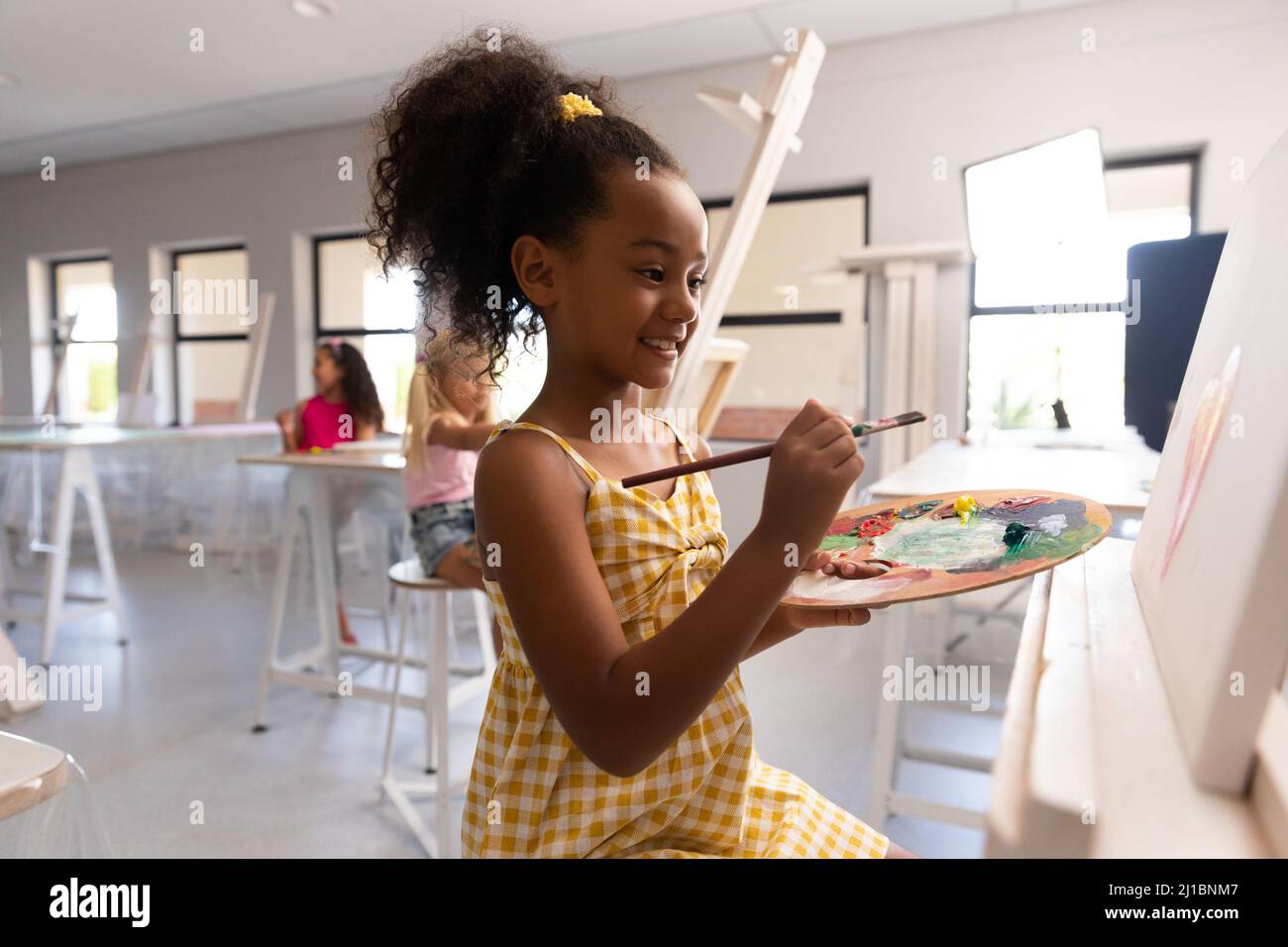 Smiling biracial elementary schoolgirl with palette and paintbrush