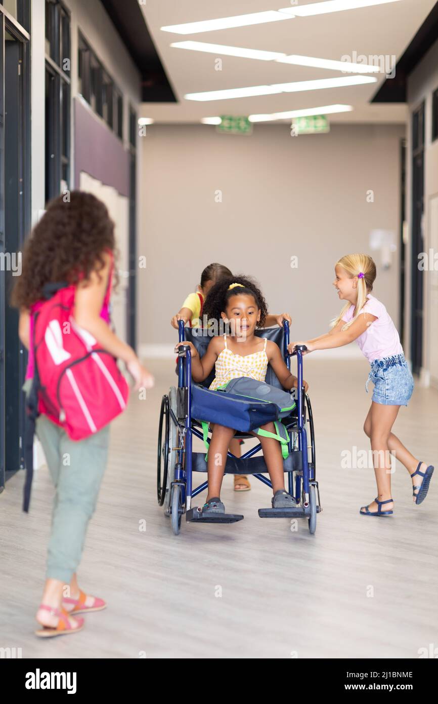 Multiracial elementary schoolgirls holding female classmate wheelchair in corridor Stock Photo