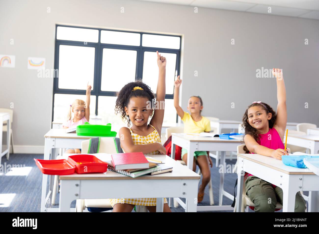 Smiling elementary schoolgirls raising hands while sitting at desk ...
