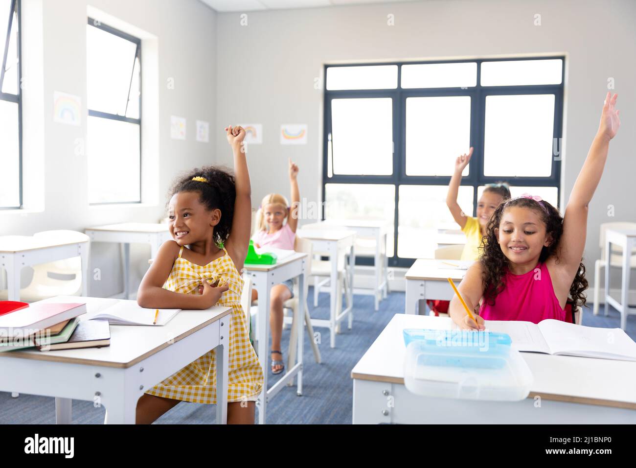 Cheerful elementary schoolgirls raising hands while sitting at desk ...
