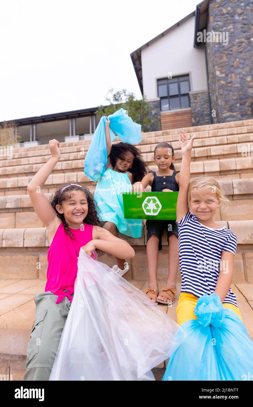 Happy multiracial elementary schoolgirls with garbage bags sitting on school steps Stock Photo