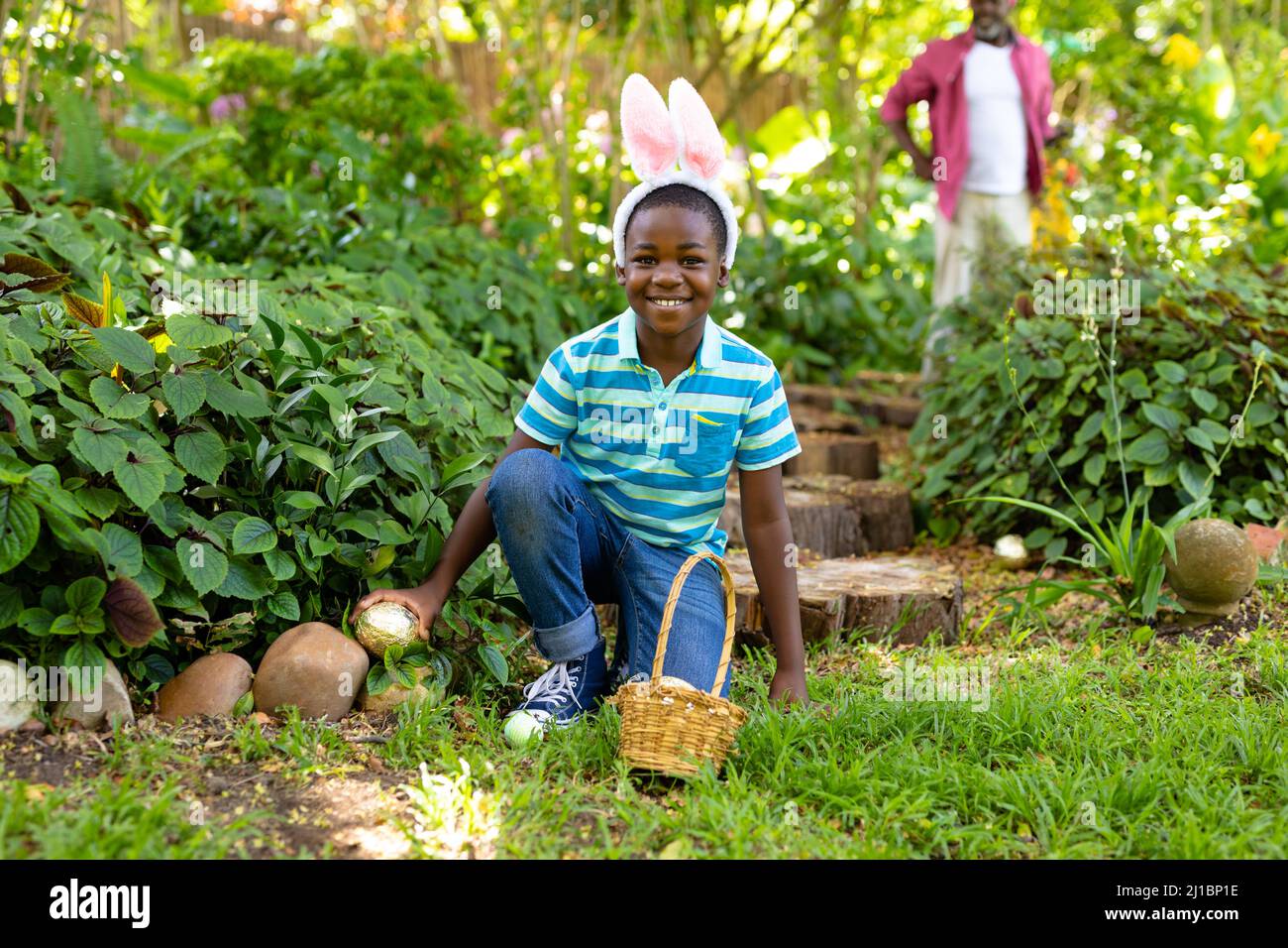 Portrait of smiling cute african american boy wearing bunny ears hiding easter egg in backyard Stock Photo