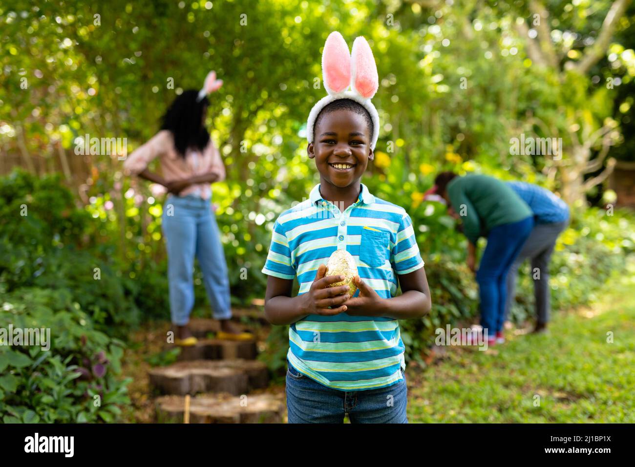 Portrait of happy african american boy in bunny ears holding easter egg while family in backyard Stock Photo