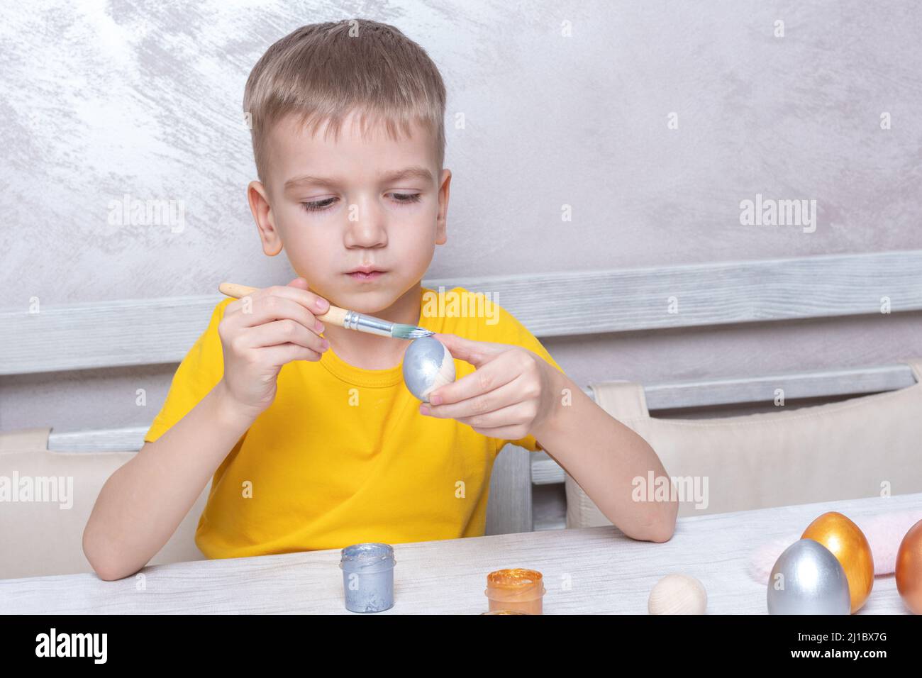A little blond boy paints eggs for the Easter holiday in the home kitchen. The child has fun and celebrates the holiday. DIY Easter eggs concept. Stock Photo